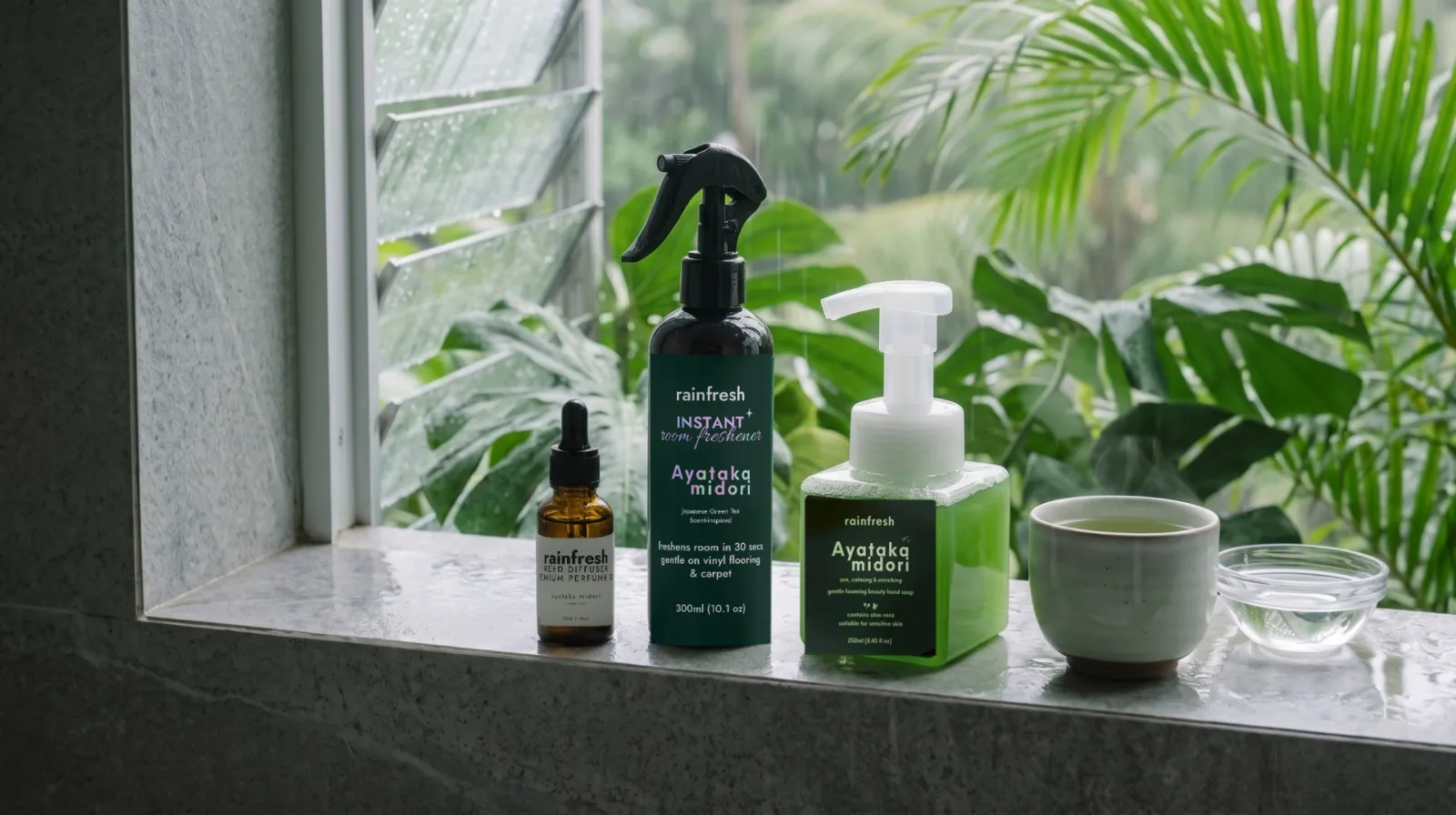 Ayataka Midori perfume oil, room freshener and hand soap on a wet stone sill beside a cup of green tea, tropical leaves and rain outside