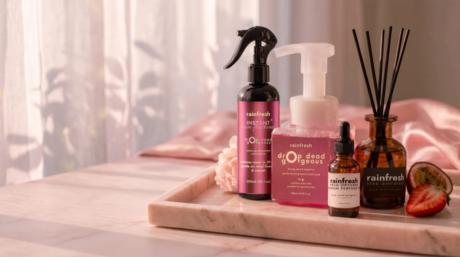 Drop Dead Gorgeous collection — room freshener, hand soap, perfume oil and reed diffuser — on a marble tray with a peony, passionfruit and strawberry, sheer curtains behind