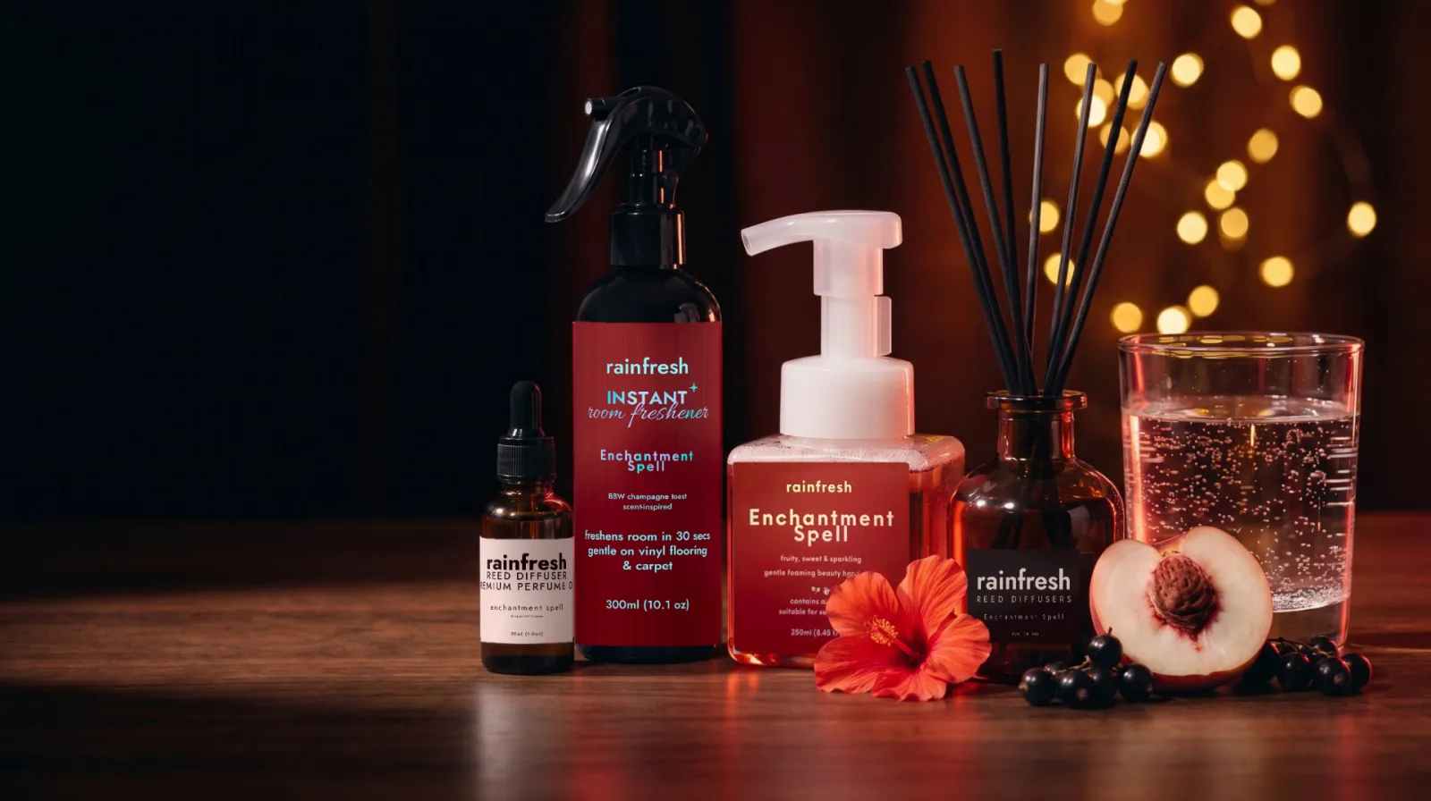 Enchantment Spell collection — perfume oil, room freshener, hand soap and reed diffuser — with a red hibiscus, white nectarine, blackcurrants and a glass of sparkling water, fairy lights behind