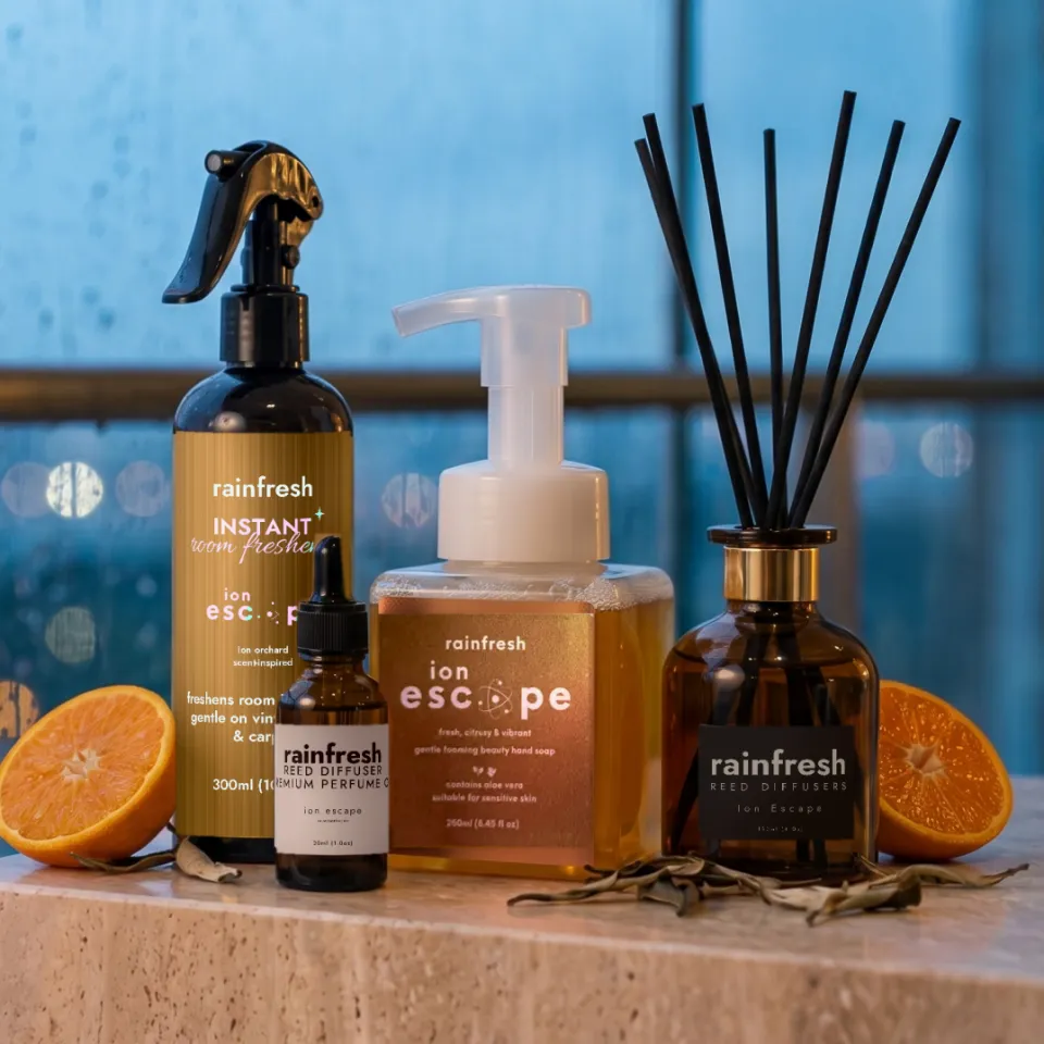 Ion Escape collection — room freshener, perfume oil, hand soap and reed diffuser — on a stone ledge with halved oranges and dried tea leaves, rain on a window behind
