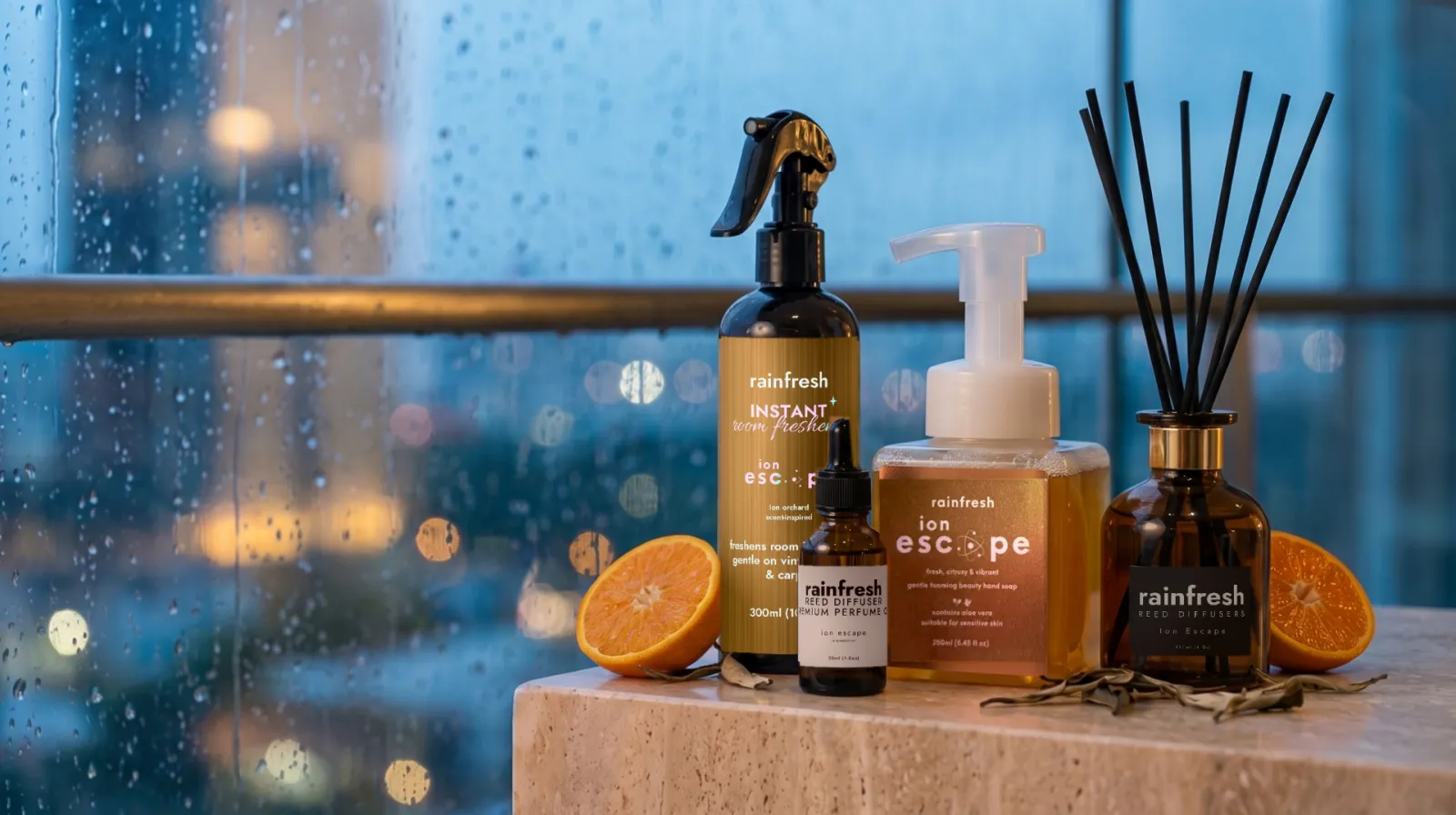 Ion Escape collection — room freshener, perfume oil, hand soap and reed diffuser — on a stone ledge with halved oranges and dried tea leaves, rain on a window behind